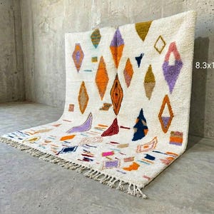 Op de afbeelding: Een witte, rechthoekige vloerkleed met een geometrisch patroon van ruiten en andere vormen in oranje, paars, blauw en bruin. Het vloerkleed meet ongeveer 253 x 314 cm en heeft een gefranjerde rand.