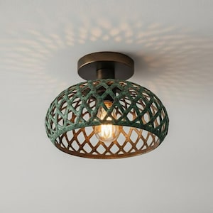 Puede incluir: Una lámpara de techo con base de bronce y pantalla calada verde. La pantalla tiene un patrón de diamantes y la bombilla es visible. La lámpara proyecta una sombra estampada en el techo.