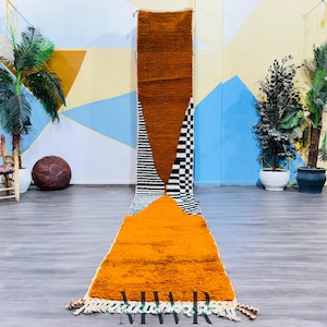 May include: A long, rectangular rug with a geometric pattern in shades of orange, white, and black. The rug has a fringe border and is laid out on a gray floor.
