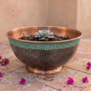 Puede incluir: Un cuenco redondo de cobre martillado con una banda horizontal ancha de pátina verde cardenillo alrededor del exterior superior. El cuenco cuenta con una base de pedestal y contiene tierra oscura con una planta suculenta verde centrada.