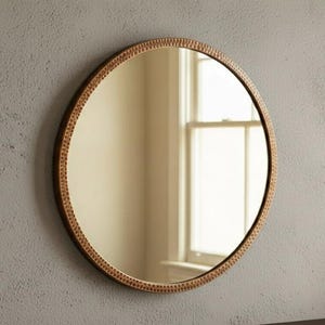 Op de afbeelding: Een ronde spiegel met een decoratieve, getextureerde koperkleurige lijst. De spiegel hangt aan een lichtgrijze muur. De weerspiegeling toont een raam met natuurlijk licht. Een kleine potplant staat eronder.