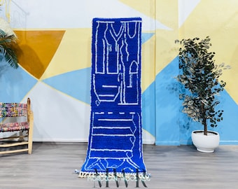 May include: A vibrant blue Moroccan rug with a white geometric design. The rug is rectangular and features a central pattern of abstract shapes. The rug is displayed vertically, showcasing its intricate details and texture. The rug has a fringe at the bottom.