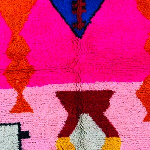 May include: A colorful rug with geometric patterns in shades of pink, orange, red, blue, and white. The rug is made of a soft, fluffy material.
