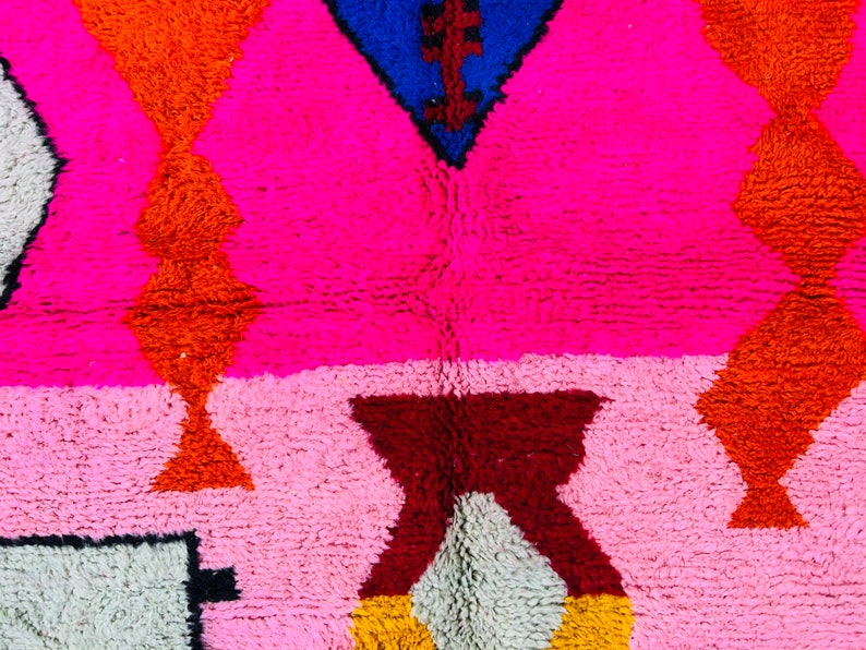 May include: A colorful rug with geometric patterns in shades of pink, orange, red, blue, and white. The rug is made of a soft, fluffy material.