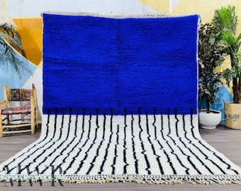Può includere: Un tappeto marocchino blu e bianco con una metà superiore blu tinta unita e una metà inferiore bianca con strisce nere. Il tappeto ha frange su tutti i lati.