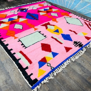 May include: A pink Moroccan rug with a geometric pattern in shades of red, blue, yellow, orange, and white. The rug has a fringe border.