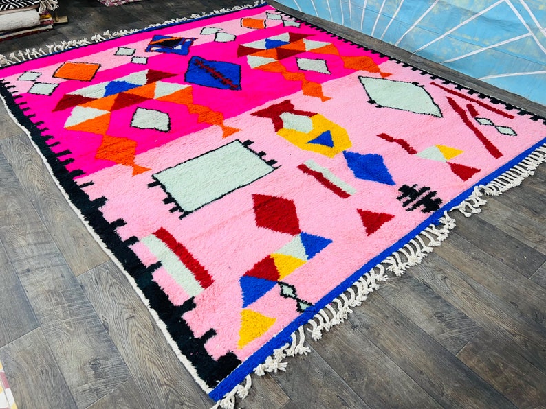May include: A pink Moroccan rug with a geometric pattern in shades of red, blue, yellow, orange, and white. The rug has a fringe border.