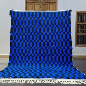 May include: A large, rectangular rug with a checkered pattern in shades of blue and black. The rug has a white fringe along the bottom edge. The rug is displayed against a white wall, with wooden windows and doors in the background.