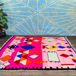 May include: A pink, red, blue, orange, and white Moroccan rug with a geometric pattern. The rug has a black border and fringe.