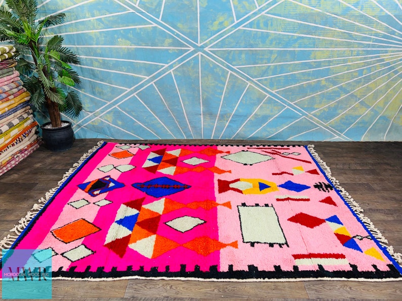May include: A pink, red, blue, orange, and white Moroccan rug with a geometric pattern. The rug has a black border and fringe.