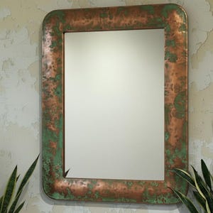 May include: Rectangular mirror with a copper-coloured frame featuring green patina accents. The mirror is set against a textured, off-white wall, with green and yellow plants in the foreground. The frame has rounded corners.