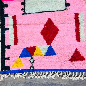 May include: A pink rug with a geometric pattern in red, blue, yellow, and white. The rug has a fringe border.