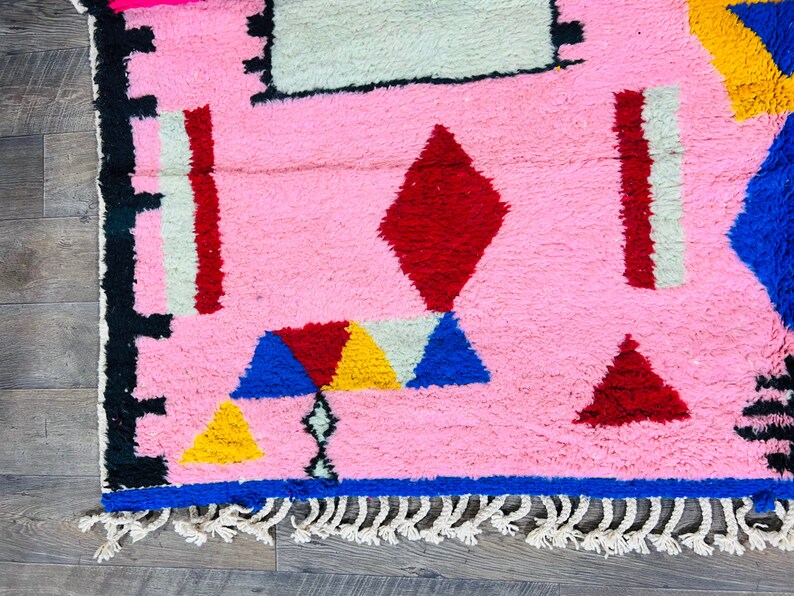 May include: A pink rug with a geometric pattern in red, blue, yellow, and white. The rug has a fringe border.