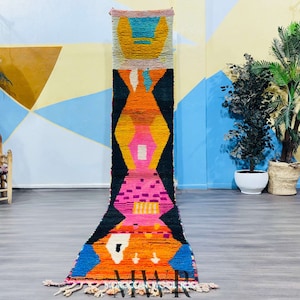 Puede incluir: Una alfombra marroquí larga y colorida con patrones geométricos en tonos de rosa, naranja, azul y negro. La alfombra está colgada en una pared en una habitación con un fondo azul claro y amarillo.