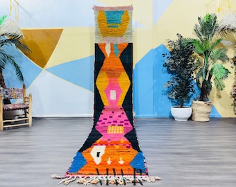 May include: A long, colorful Moroccan rug with geometric patterns in shades of pink, orange, blue, and black. The rug is hanging on a wall in a room with a light blue and yellow background.