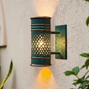 Puede incluir: Aplique de pared para exteriores verde y bronce con una pantalla cilíndrica estampada. La lámpara está montada en una pared beige texturizada, con follaje en primer plano. La lámpara emite un brillo cálido y acogedor.