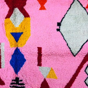 May include: A pink rug with a geometric pattern in shades of red, yellow, blue, white, and black. The rug features a variety of shapes, including diamonds, triangles, and rectangles.