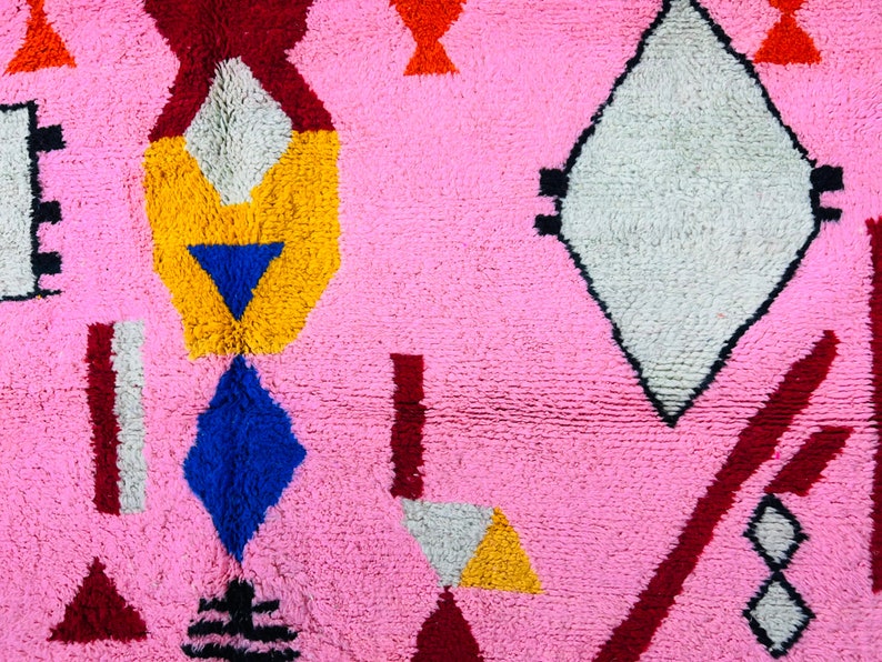 May include: A pink rug with a geometric pattern in shades of red, yellow, blue, white, and black. The rug features a variety of shapes, including diamonds, triangles, and rectangles.