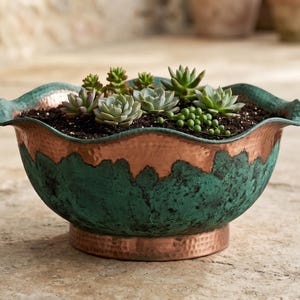 Pode incluir: Um vaso em forma de taça com borda recortada, apresentando um acabamento em cobre martelado com uma pátina texturizada em tom verde-azulado escuro. A taça está preenchida com terra escura e várias pequenas suculentas verdes de formas variadas.