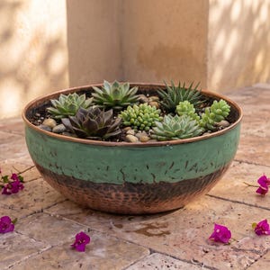 May include: A round, shallow bowl with a two-tone finish featuring a matte green upper section and a hammered copper base. The bowl is filled with a variety of small green and dark purple succulents planted in soil with small pebbles.
