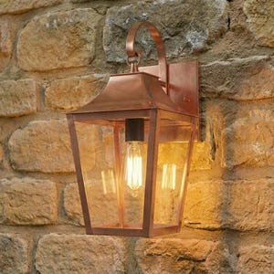 May include: A copper outdoor wall lantern with a clear glass enclosure and a decorative bulb. The lantern is mounted on a weathered stone wall. The lantern's design features a handle at the top.