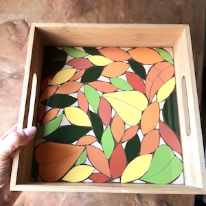 Puede incluir: Una bandeja de servir de madera con un diseño de mosaico de azulejos en varios colores otoñales, incluyendo naranja, amarillo, verde y rojo. Los azulejos están dispuestos en un patrón de hojas.