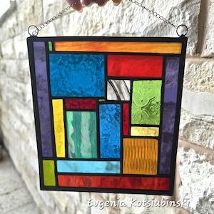 Op de afbeelding: Een glas-in-loodpaneel met een zwarte rand, met een geometrisch ontwerp van kleurrijke glazen stukken. Het paneel bevat blauwe, rode, groene, gele en oranje secties. Het glas heeft textuur en patronen en wordt opgehangen aan een ketting.