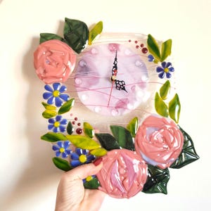 May include: A pink and white glass clock with a floral design. The clock face is surrounded by pink roses, blue flowers, and green leaves.