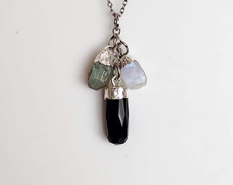 Könnte beinhalten: Eine silberfarbene Kettenhalskette mit einem Bündel aus drei Anhängern. Die Charms bestehen aus einem facettierten schwarzen zylindrischen Stein, einem rohen grünen Stein und einem rohen schillernden weißen Stein, jeweils mit silberfarbenen Metallkappen.