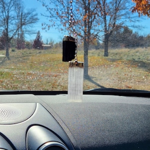 May include: A car charm featuring a black rectangular stone and a clear, cylindrical crystal pendant, both with gold-colored metal accents. The charm hangs from a silver chain. The background shows a sunny day with trees and a grassy field.