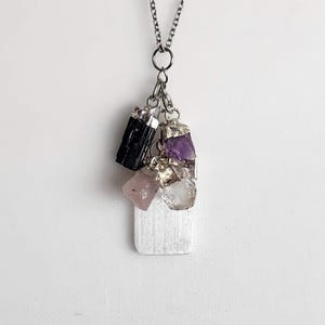 May include: A silver-toned necklace featuring a cluster of raw gemstones. The pendant includes a black tourmaline, amethyst, rose quartz, and clear quartz, all set in silver. A rectangular, textured selenite crystal hangs at the bottom.