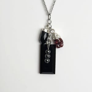 May include: A silver chain necklace with a pendant featuring a black rectangular onyx stone, three small silver beads, a faceted black stone, and a raw red garnet gemstone. The necklace is displayed against a white background.