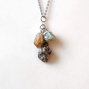 May include: A silver-toned necklace featuring three natural stone pendants. The stones include a golden-brown citrine, a pale blue-green aquamarine, and a textured, metallic pyrite. The necklace hangs against a white background.