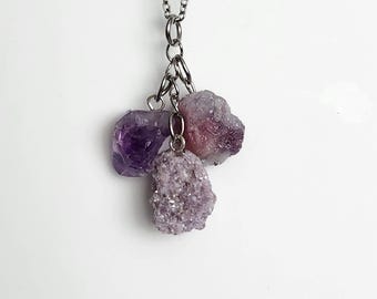 May include: A silver-toned metal chain necklace featuring three dangling raw gemstone pendants. One pendant is a smooth, deep purple amethyst, while the other two are textured, light lavender-colored druzy crystal clusters hanging from metal rings.