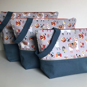 Könnte beinhalten: Drei Tragetaschen mit grauem Hintergrund und einem farbenfrohen Lama- und Blumenmuster. Die Taschen haben einen blauen Boden und Griffe, mit einem goldenen Reißverschluss und Besatz. Die Taschen sind in verschiedenen Größen.