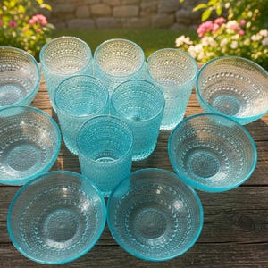May include: A collection of clear, turquoise-colored glassware, including bowls and tumblers, arranged on a weathered wooden table. The glassware features a textured, dotted pattern. The scene is set outdoors with flowers in the background.