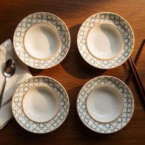 May include: Four white ceramic bowls with a beige rim and a repeating black circle pattern. A spoon and a napkin are on the left, and chopsticks are on the right. The bowls are on a dark wooden surface.