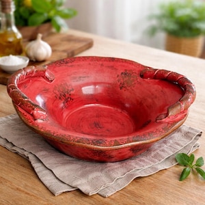 May include: A rustic, red ceramic bowl with brown accents and rope-like handles. The bowl sits on a neutral-colored linen napkin on a wooden table. The bowl's interior is a vibrant red, and the exterior has a distressed finish.