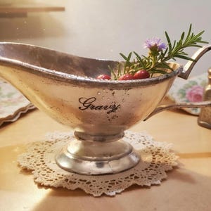 May include: Silver gravy boat with the word "Gravy" in script. The boat is filled with rosemary, small purple flowers, and red berries. It sits on a lace doily. A small salt shaker and floral napkin are in the background.