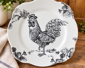 May include: A white ceramic plate with a scalloped edge featuring a black line-art illustration of a rooster standing in grass, surrounded by botanical accents including leafy vegetables and foliage around the perimeter of the plate.