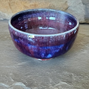 May include: A handmade ceramic bowl with a glossy, iridescent finish. The bowl features a blend of deep purple, blue, and maroon hues. The rim has a lighter, almost white, edge. The bowl is round and sits on a dark surface.