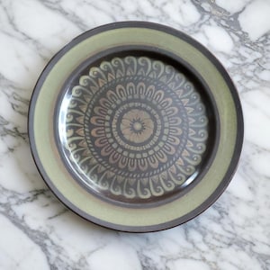 May include: A round ceramic plate with a decorative mandala pattern. The plate features a central floral design surrounded by concentric circles of repeating patterns in shades of brown, green, and gray. The rim has a green border.