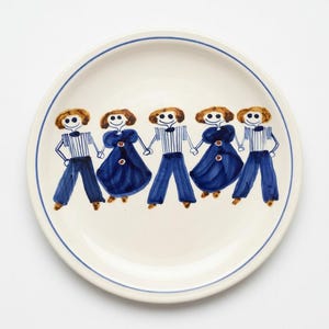 May include: A white ceramic plate with a blue rim and a whimsical hand-painted design. The design features five stylized figures in blue and white outfits, holding hands. The figures have brown hair and are smiling. A charming decorative plate.