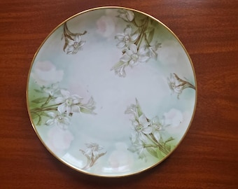 May include: A round porcelain plate featuring a pale green and white background with hand-painted white daffodil flowers and green stems scattered across the surface. The plate is finished with a thin gold trim along the circular edge.