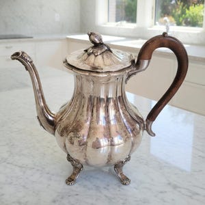 May include: A silver-colored antique coffee pot with a wooden handle and a decorative lid. The pot has a curved spout and sits on three ornate feet. The lid is topped with a small, decorative bird.