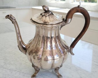 May include: A silver-colored antique coffee pot with a wooden handle and a decorative lid. The pot has a curved spout and sits on three ornate feet. The lid is topped with a small, decorative bird.