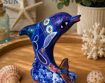 May include: A blue ceramic dolphin figurine with floral and geometric patterns. The dolphin has a red mouth and is set on a blue wave base. Seashells and a decorative sign with the text "SEA SALT & SUN" are in the background.