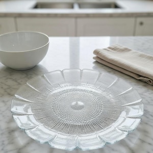 May include: A clear glass plate with a floral design, featuring a central textured circle and radiating lines. The plate is on a marble surface, with a white bowl and folded beige cloth nearby. Kitchenware.