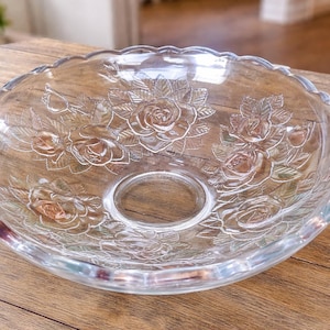 May include: Clear glass serving bowl with a scalloped edge and a central opening. The bowl features an embossed rose pattern. The bowl is sitting on a wooden surface.