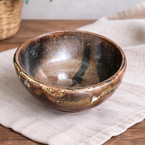 May include: A handmade ceramic bowl with a speckled, glazed finish. The bowl features a dark brown interior with black and tan accents, and a lighter brown rim. It sits on a beige linen napkin on a wooden surface.